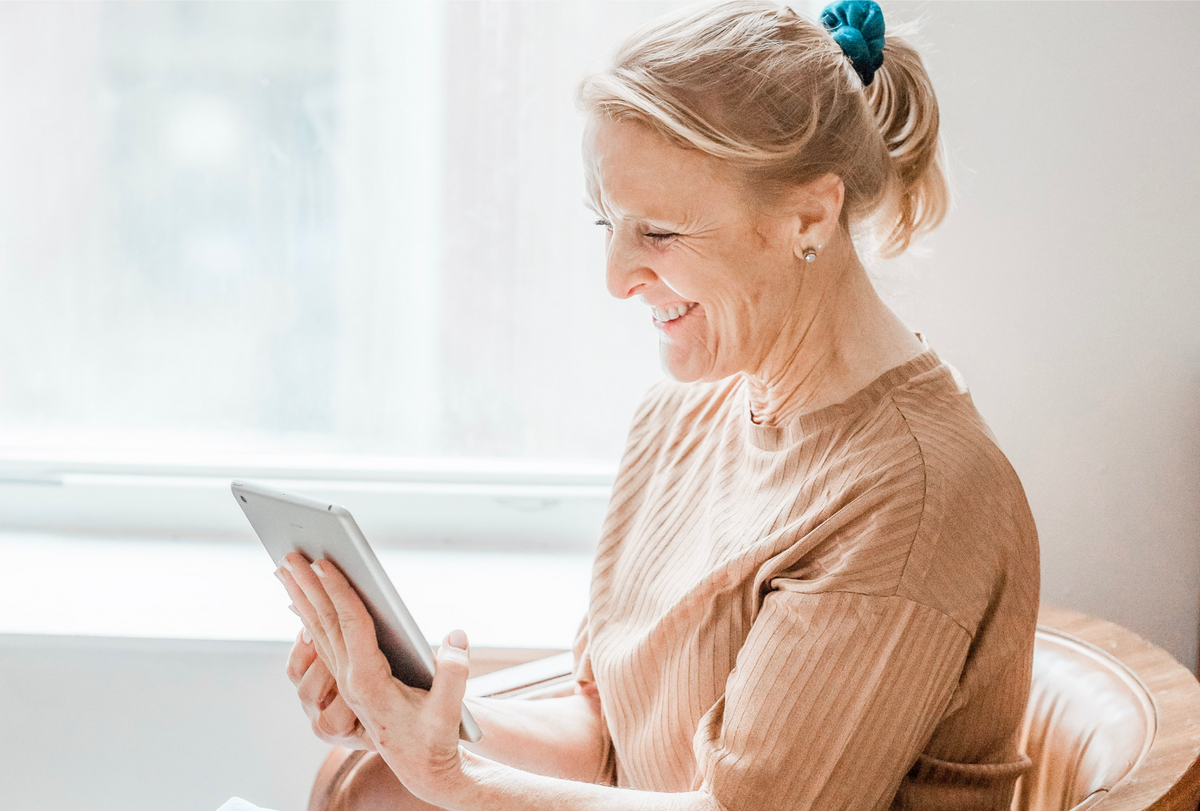 Older woman sitting by a bright window, smiling while using a tablet