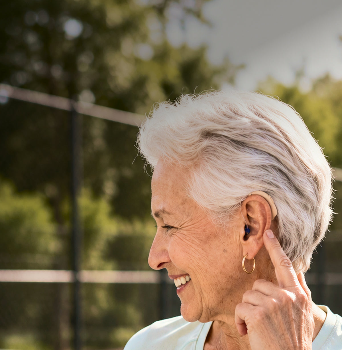 An older woman with short white hair is shown in profile outdoors, touching her ear where a small dark in-ear device is placed. She is wearing a light-colored top and a gold hoop earring, with a blurred green outdoor background.