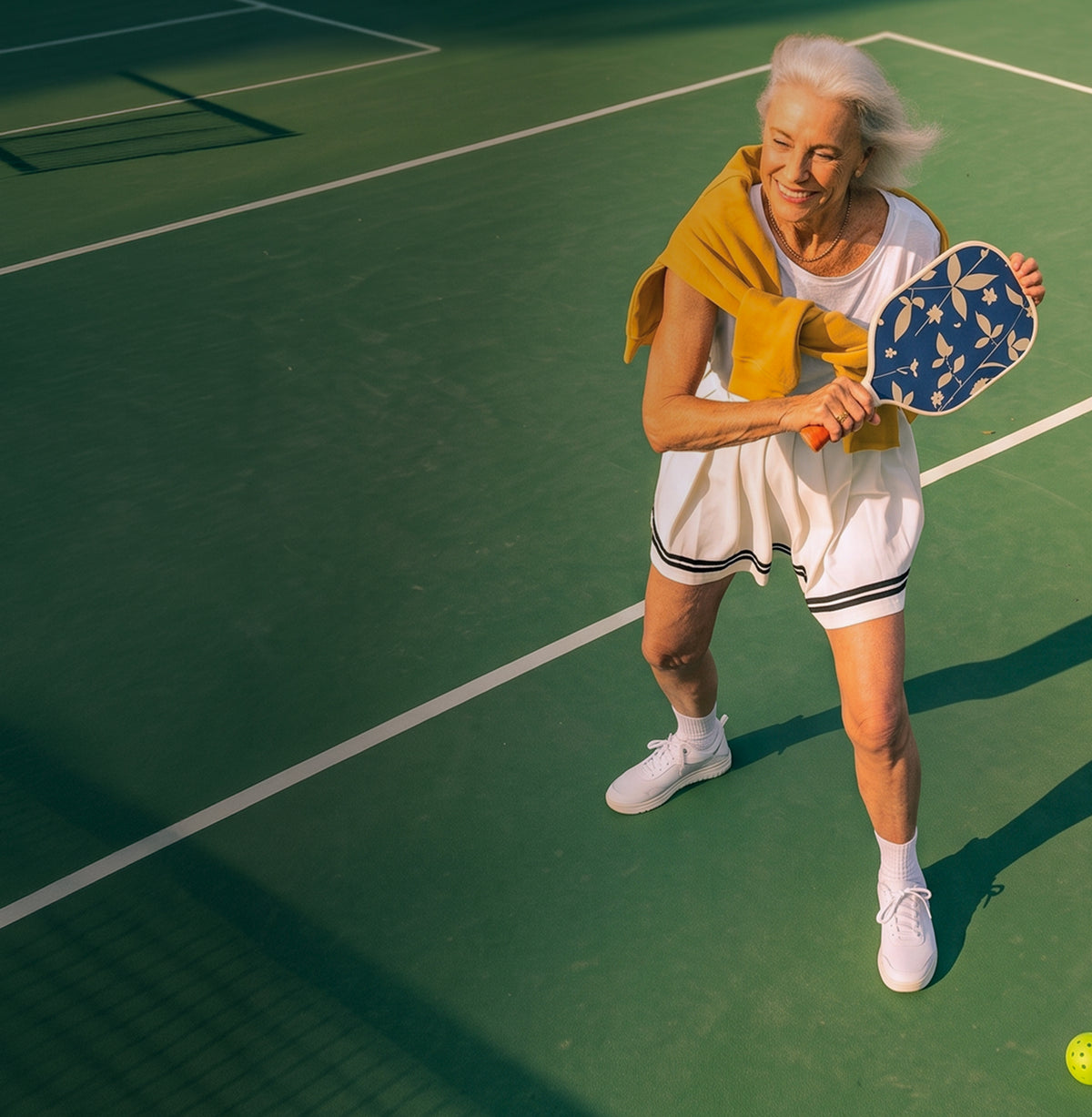 An older woman playing pickleball on a green court, wearing a white athletic dress with black trim, white sneakers, and holding a blue paddle with a yellow towel draped over her shoulder.